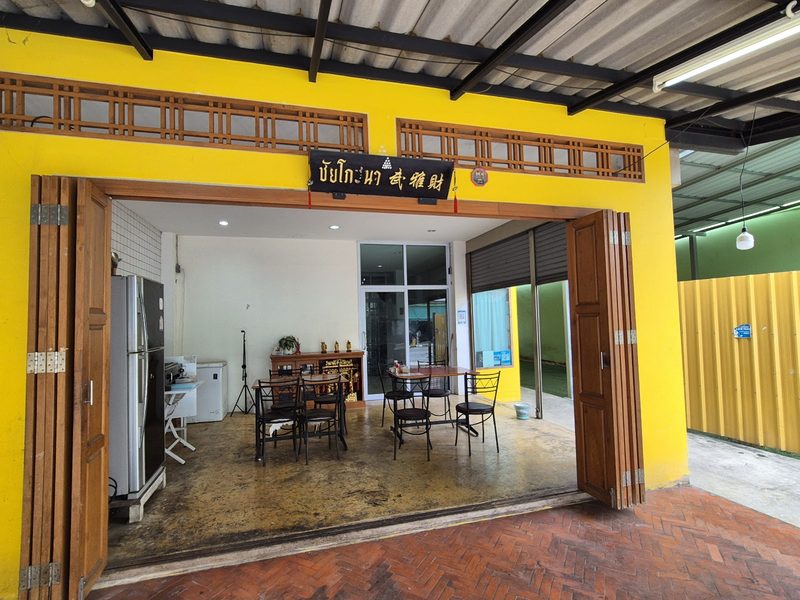 หน้าร้านชัยโภชนา ผนังสีเหลือง ประตูไม้บานเฟี้ยมเปิดเห็นโต๊ะในห้อง และป้ายชื่อร้านสีดำเหนือประตู